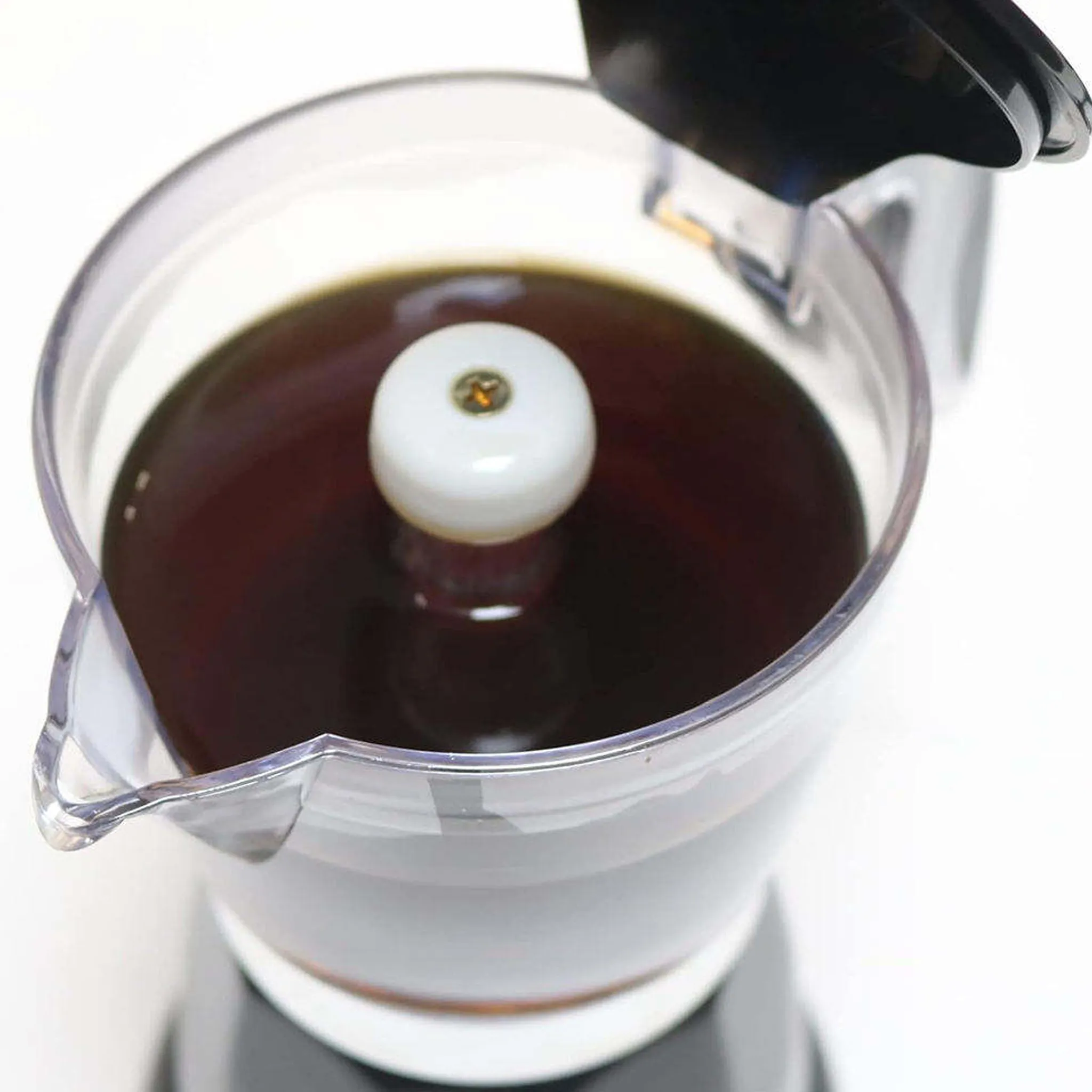Primer plano de la jarra transparente de una cafetera moka llena de café recién hecho, con la tapa negra abierta y la columna central blanca visible, sobre fondo blanco.