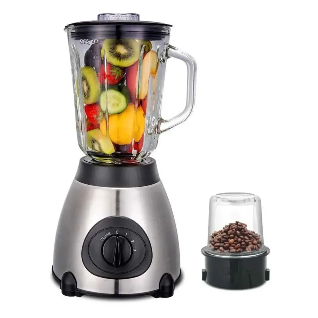 Licuadora de acero inoxidable con jarra de vidrio llena de frutas y verduras, junto a un accesorio molinillo pequeño con granos de café, aislada sobre fondo blanco.